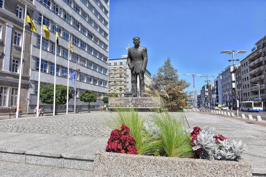 Na pierwszym planie: betonowa donica z roślinami. Na drugim planie: bryła pomnika kaszubskiego działacza. W tle miejska zabudowa.