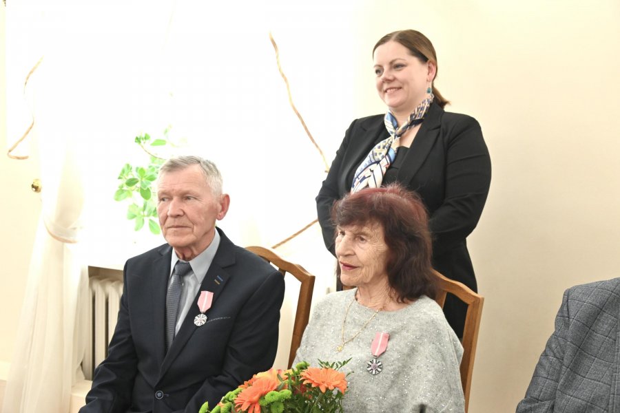 Para starszych os&oacute;b, pani z bukietem kwiat&oacute;w, z tyłu kobieta w granatowej marynarce