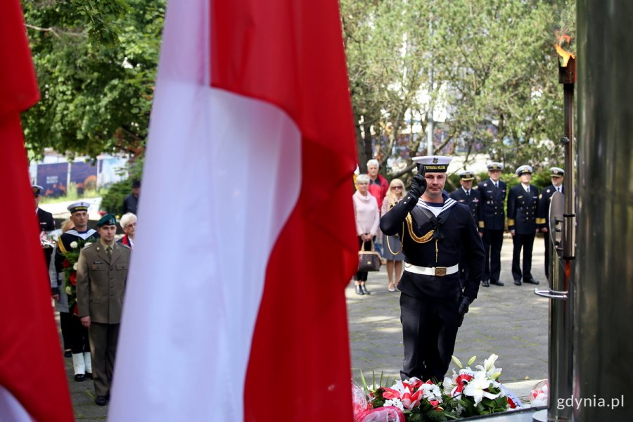 Gdyńskie obchody 82. rocznicy agresji niemieckiej na Polskę. Na pierwszy planie biało-czerwona flaga. Obok niej widać marynarza, który salutuje przed pomnikiem Obrońców Wybrzeża na Cmentarzu Wojennym w Redłowie. W tle inni uczestnicy obchodów // fot. Przemysław Kozłowski