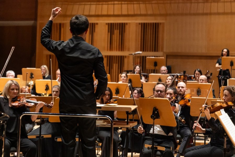Koncert najlepszych dyplomantów Akademii Muzycznej im. Stanisława Moniuszki w Gdańsku podczas gali Pomorskiej Nagrody Artystycznej. Na pierwszym planie dyrygent w uniesioną do góry ręką. Za nim muzycy grający na instrumentach. Stoją przed nimi pulpity z nutami