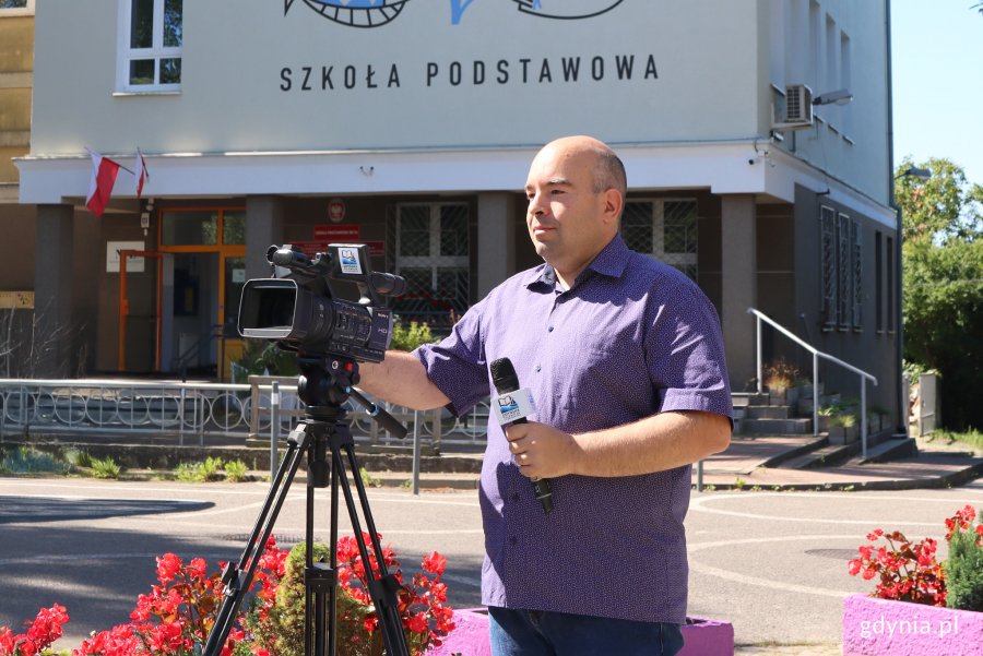 Upalny dzień, mężczyzna przygotowuje kamerę video do nagrania, w tle budynek Zespolu Szkół Ogólnokształcących nr 6 w Gdyni z grafiti na fasadzie