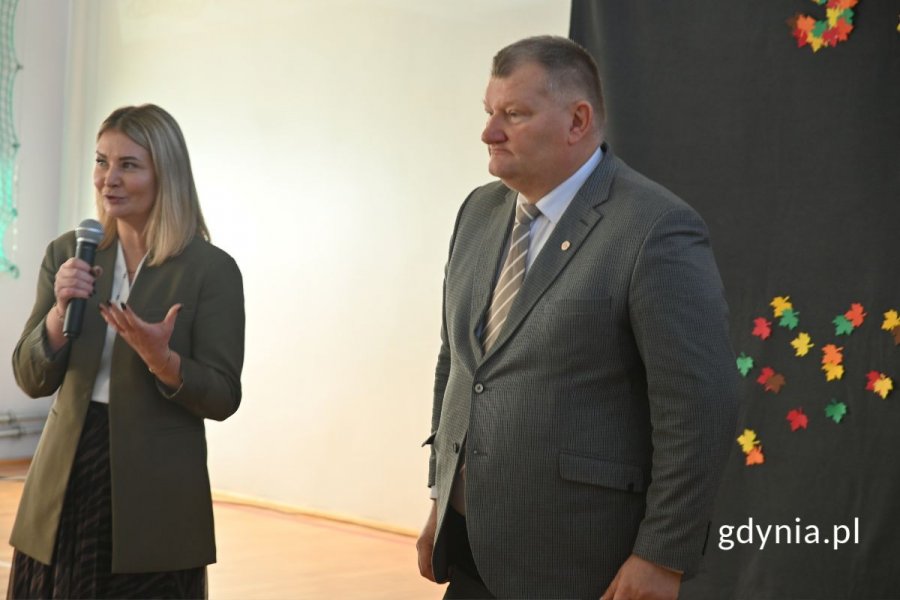 Stojąca kobieta mówiąca do mikrofonu w szkolnej hali sportowej i stojący obok mężczyzna