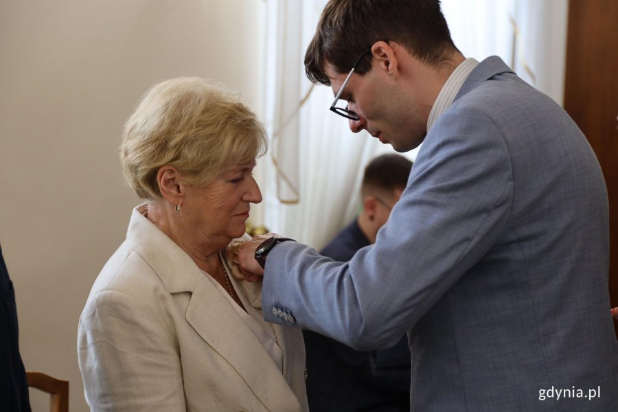 Mężczyzna w garniuturze przypina medal starszej pani w białej marynarce
