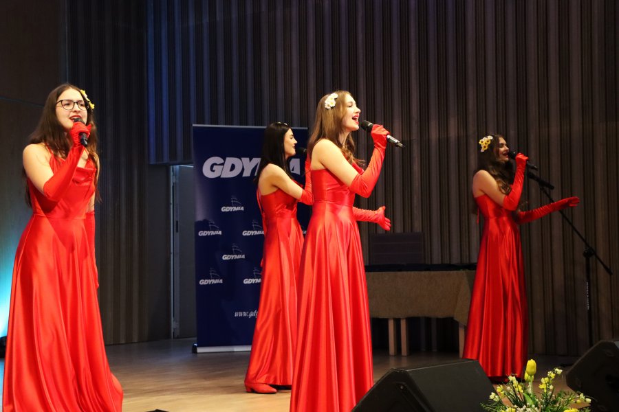 Cztery dziewczyny w czerwonych sukniach, śpiewające na scenie do mikrofonów
