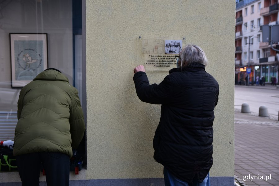 Mężczyzna po prawej trzyma przezroczystą tablicę pamiątkową z czarnymi napisami, mapką i zdjęciem budynku, w którym mieścił się pierwszy gdyński magistrat. Mężczyzna po lewej pochyla się nad skrzynką z narzędziami // fot. Sławomir Okoń