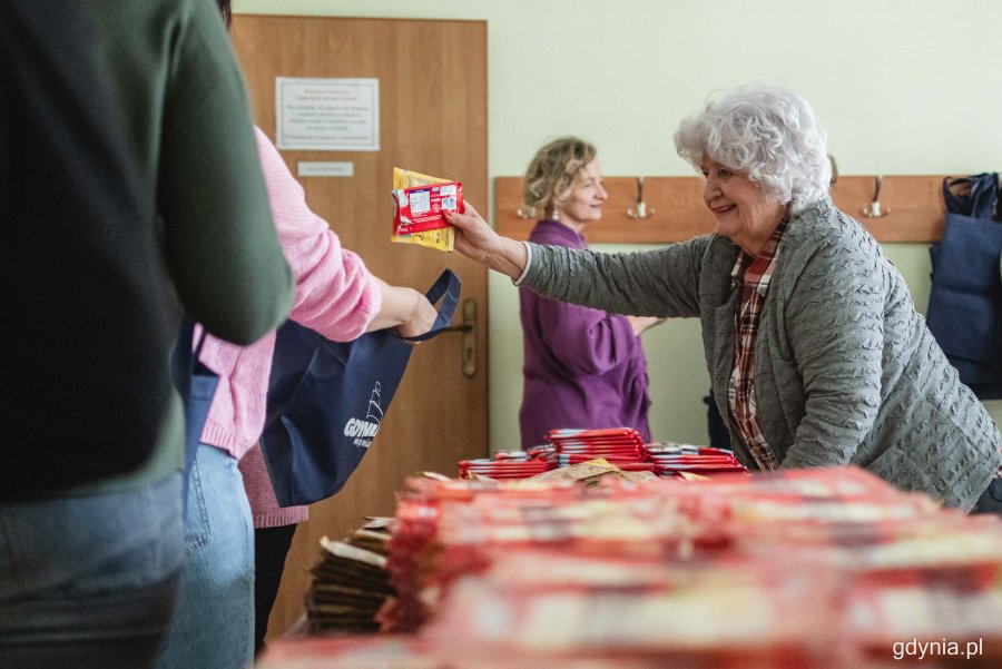 Pakowanie wielkanocnych paczek dla potrzebujących, które odbyło się w Centrum Aktywności Seniora. Uśmiechnięta seniorka wrzuca produkty spożywcze do torby, którą trzyma inna wolontariuszka // fot. Kamil Złoch