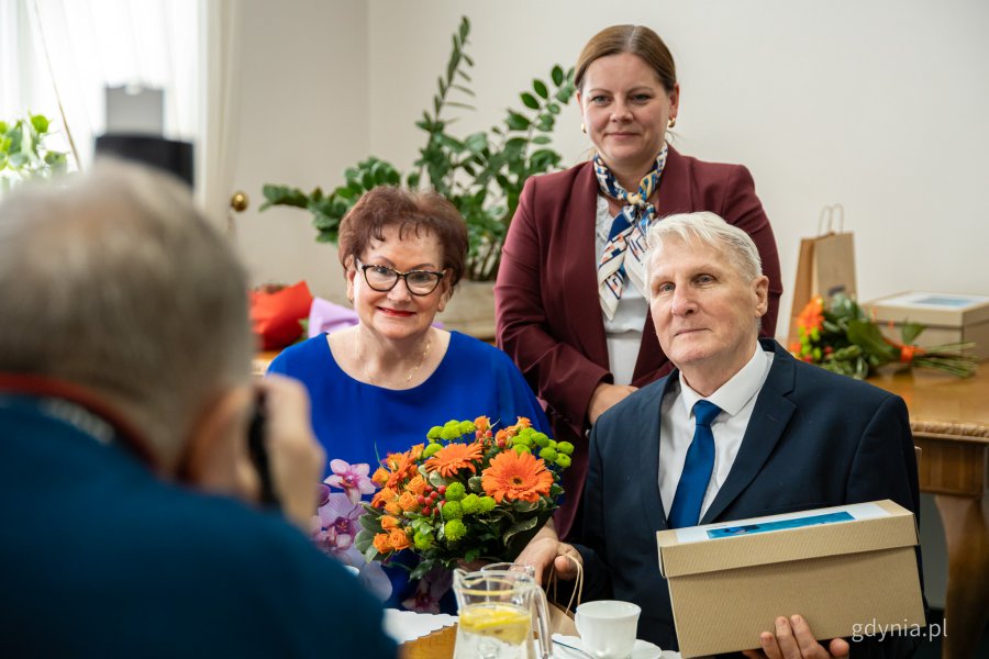 Prezydent Gdyni Aleksandra Kosiorek stojąca za plecami siedzącej przy stole pary małżeńskiej
