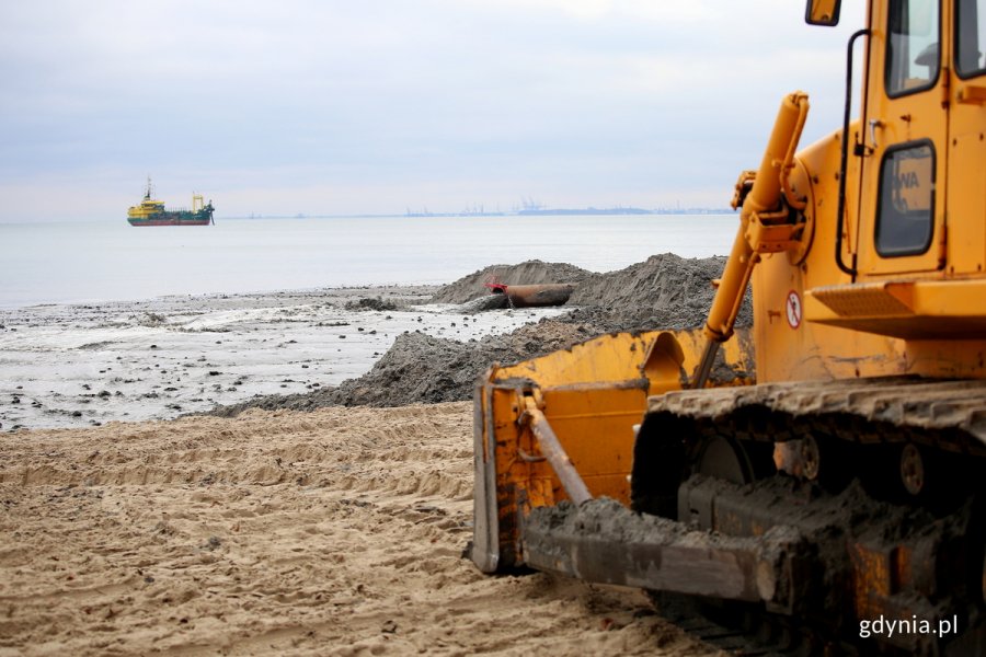 Woda płynie z rury na plaży, na pierwszym planie koparka.