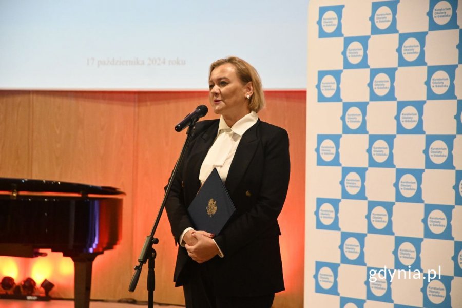 Kobieta elegancko ubrana i trzymająca teczkę stoi na scenie przed mikrofonem