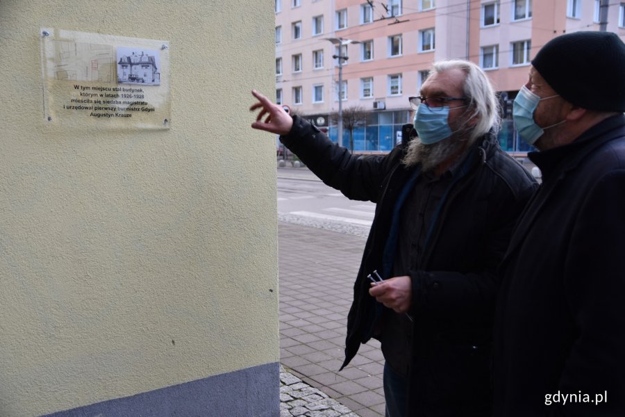 Marek Stępa, naczelnik Wydziału Ochrony Dziedzictwa Urzędu Miasta Gdyni (po prawej) przygląda się montażowi tablicy pamiątkowej, która jest umieszona na żółtej fasadzie budynku na rogu ulic Świętojańskiej i Derdowskiego. Jeden z mężczyzn pracujących przy montażu wskazuje na tablicę // fot. Sławomir Okoń