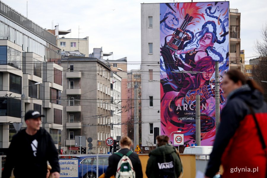 Budynki przy ulicy Partyzantów. Na jednym z nich mural przedstawiający bohaterki serialu „Arcane”. Na pierwszym planie przechodnie.