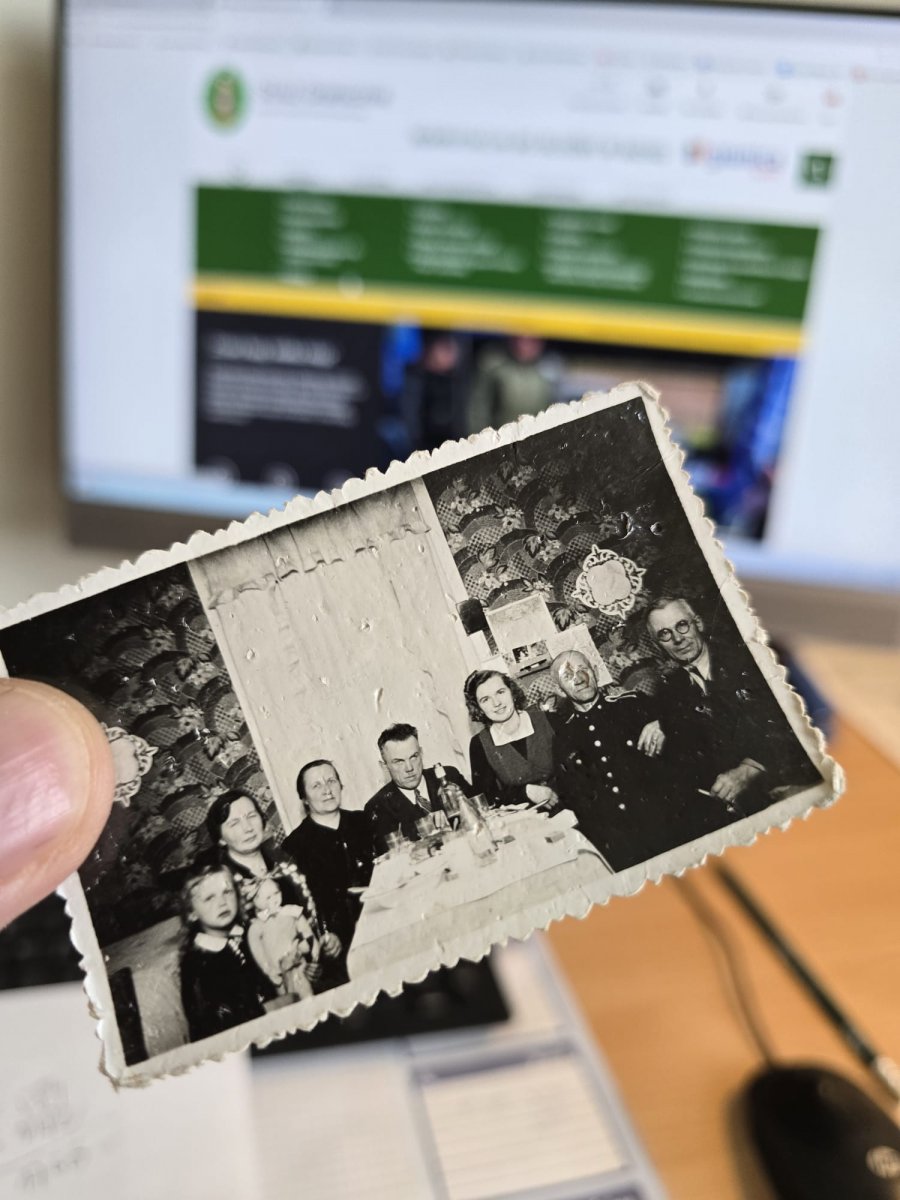 Fotografia &ndash; sądząc po stylu ubioru, fryzurach i charakterystycznym wystroju wnętrza mogła zostać wykonana w latach 40. lub 50. XX wieku. W czarno-białym kadrze ujęto kilka os&oacute;b w r&oacute;żnym wieku, w tym dziecko z lalką i mężczyznę w mundurze, siedzących przy wsp&oacute;lnym stole &ndash; zapewne rodzinę uchwyconą podczas uroczystego spotkania.