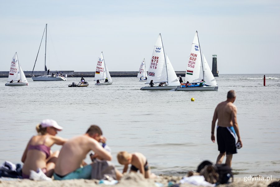 W tle płynął żaglówki, na pierwszym planie siedzą plażowicze.