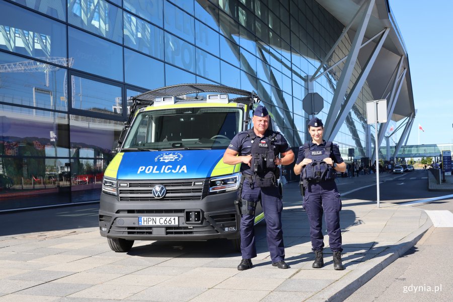 Dwie osoby z policji na tle samochodu służbowego w niebiesko-żółtym w tle budynek lotniska