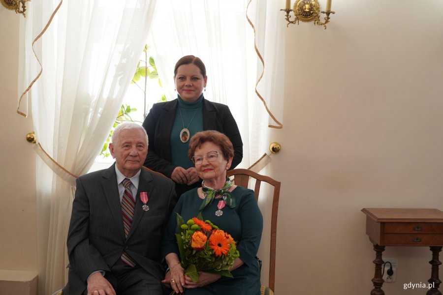 Para siedzi, kobieta trzyma pomarańczowe kwiaty, z tyłu stoi inna kobieta