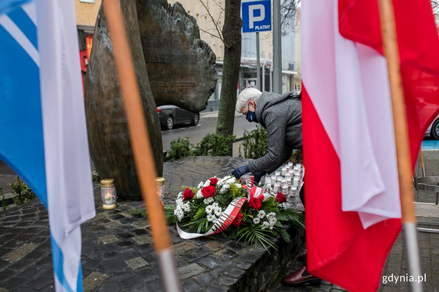 Joanna Zielińska - przewodnicząca Rady Miasta Gdyni składa wieniec z biało-czerwoną szarfą pod pomnikiem Ofiar Terroru Komunistycznego. Na pierwszym planie biało-czerwona flaga narodowa oraz biało-niebieska flaga Gdyni. Za wieńcem widać ustawione w rzędach znicze // fot. Karol Stańczak