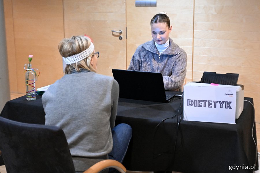 Sala w Polsat Plus Arenie Gdynia, stolik, tabliczka z napisem "Dietetyk", kobieta przy laptopie, tyłem do kadru starsza kobieta