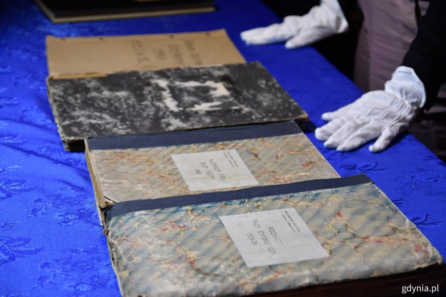 Część dokumentów dotyczących historii Marynarki Wojennej, która trafiła do Muzeum Marynarki Wojennej. tomy akt leżą na stole przykrytym niebieskim obrusem. Na zdjęciu widać dłonie w białych rękawiczkach, które układają dokumenty // fot. Marcin Mielewski