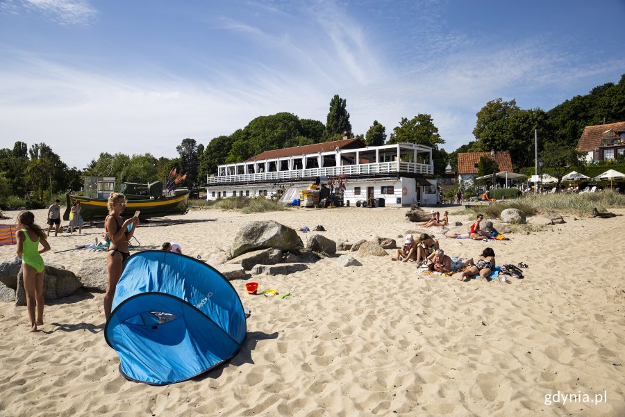 Plaża w Orłowie. Na pierwszym planie młoda kobieta w kostiumie kąpielowym stoi przy małym, niebieskim namiocie plażowym, nieco dalej leżą na piasku obok dużych kamieni inni plażowicze, w tle długi, biały pawilon rybacki i domki kryte czerwoną dachówką, za nimi zielone drzewa. Fot, Przemysław Kozłowski