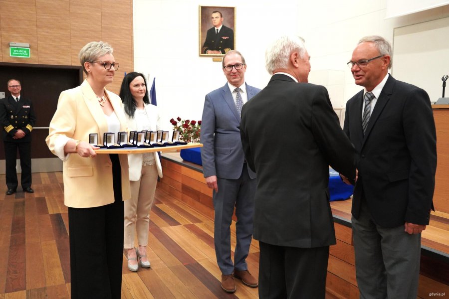 Mężczyźni w garniturach, kobieta w ż&oacute;łtej marynasrce trzyma tacę z odznaczeniami