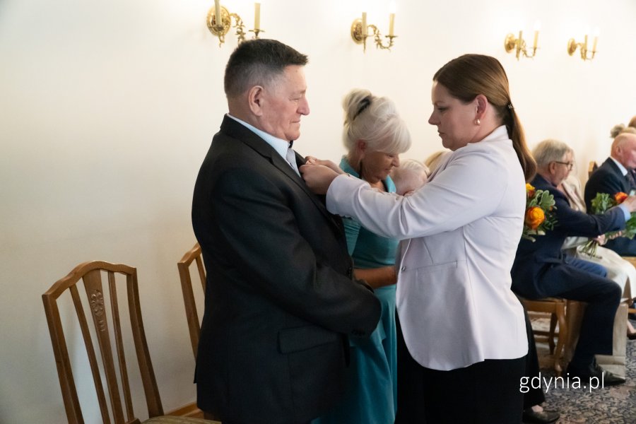 Kobieta w różowej marynarce przypina medal starszemu mężczyźnie