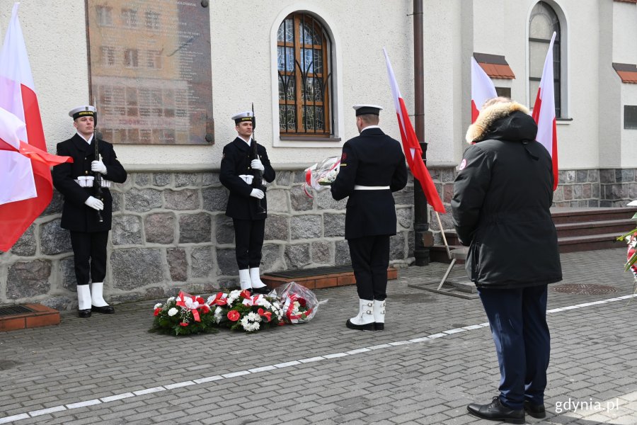 Posterunek honorowy przy tablicy upamiętniającej ofiary katastrofy smoleńskiej, która znajduje się na elewacji Bazyliki Morskiej. Jeden z marynarzy składa kwiaty pod tablicą. Obok marynarzy są stojaki z biało-czerwonymi-flagami // fot. Magdalena Czernek