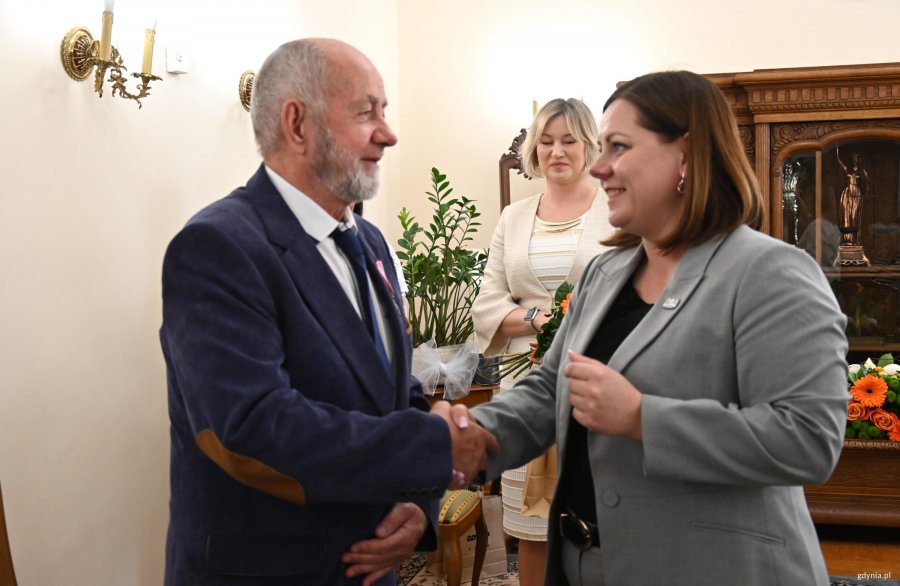 Kobieta w szarej marynarce ściska dłoń starszego mężczyzny w garniturze