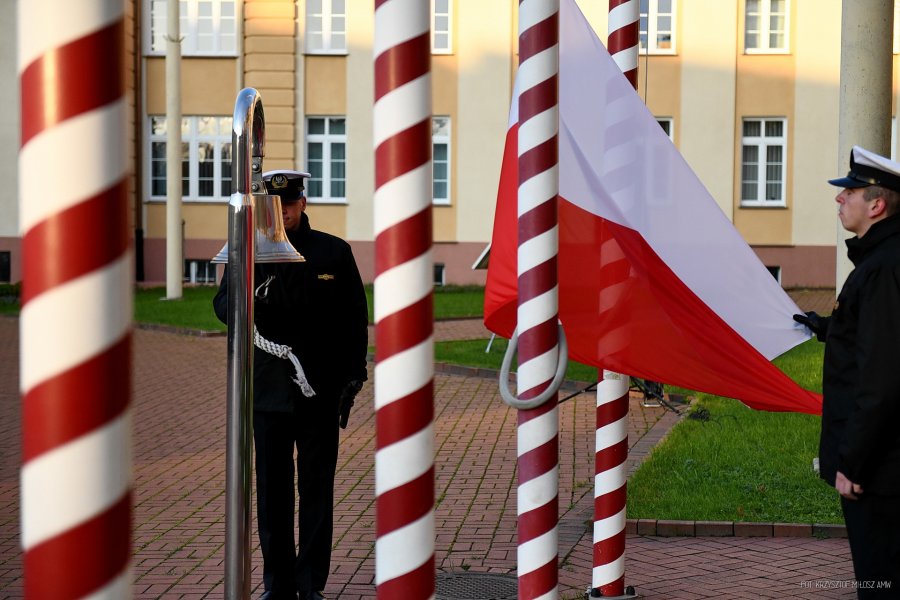 Jeden mężczyzna w marynarskim mundurze uderza w dzwon, drugi mężczyzna wciąga na maszt flagę Polski.