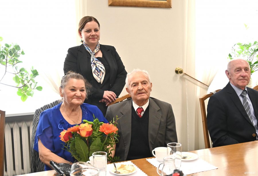 Para senior&oacute;w na krzesłach za nimi stoi kobieta w ciemnej marynarce