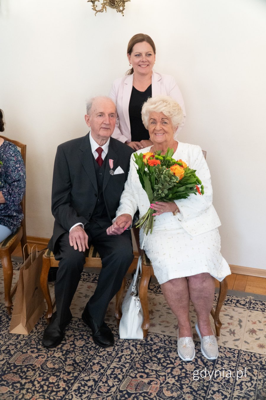 Meżczyzna w garniutrze i kobieta w eleganckim stroju z bukietem kwiatów, trzymają się za ręce