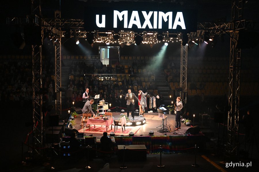Hala Polsat Plus Arena Gdynia, scena, dekoracja ze stołem i krzesłami, aktorzy na scenie, nad sceną wyświetlacz z napisem "U Maxima"