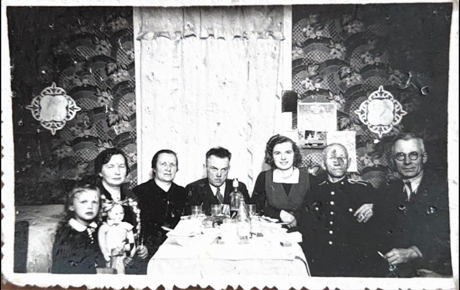 Fotografia &ndash; sądząc po stylu ubioru, fryzurach i charakterystycznym wystroju wnętrza mogła zostać wykonana w latach 40. lub 50. XX wieku. W czarno-białym kadrze ujęto kilka os&oacute;b w r&oacute;żnym wieku, w tym dziecko z lalką i mężczyznę w mundurze, siedzących przy wsp&oacute;lnym stole &ndash; zapewne rodzinę uchwyconą podczas uroczystego spotkania.