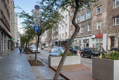 Ulica Starowiejska, brukowana jezdnia, zaparkowane auta, kamienice, na pierwszym planie podest z desek i drzewo