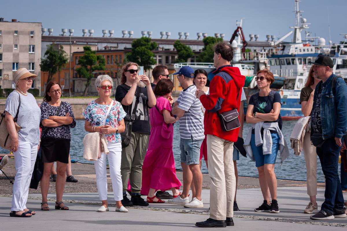 grupa os&oacute;b stojących przy nabrzeżu, w tle mająca budynki i statki