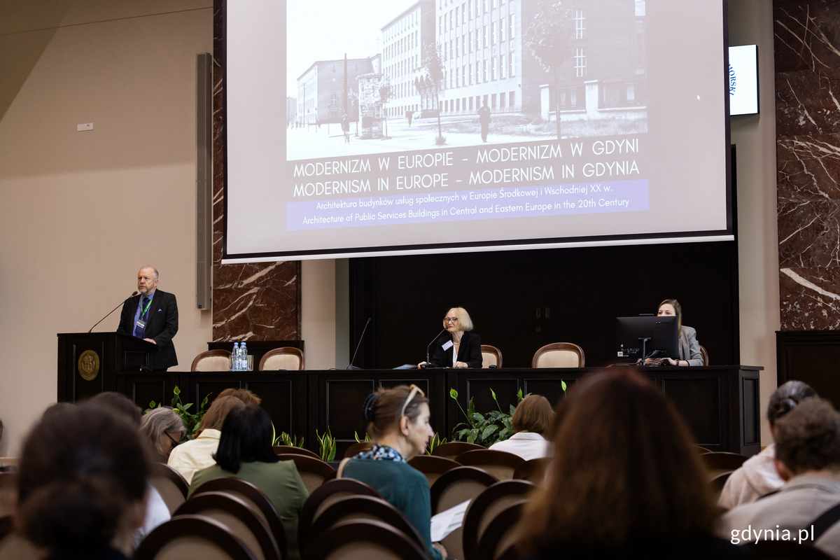 Trzy osoby na podjum w auli, na kt&oacute;rej siedzą słuchacze.