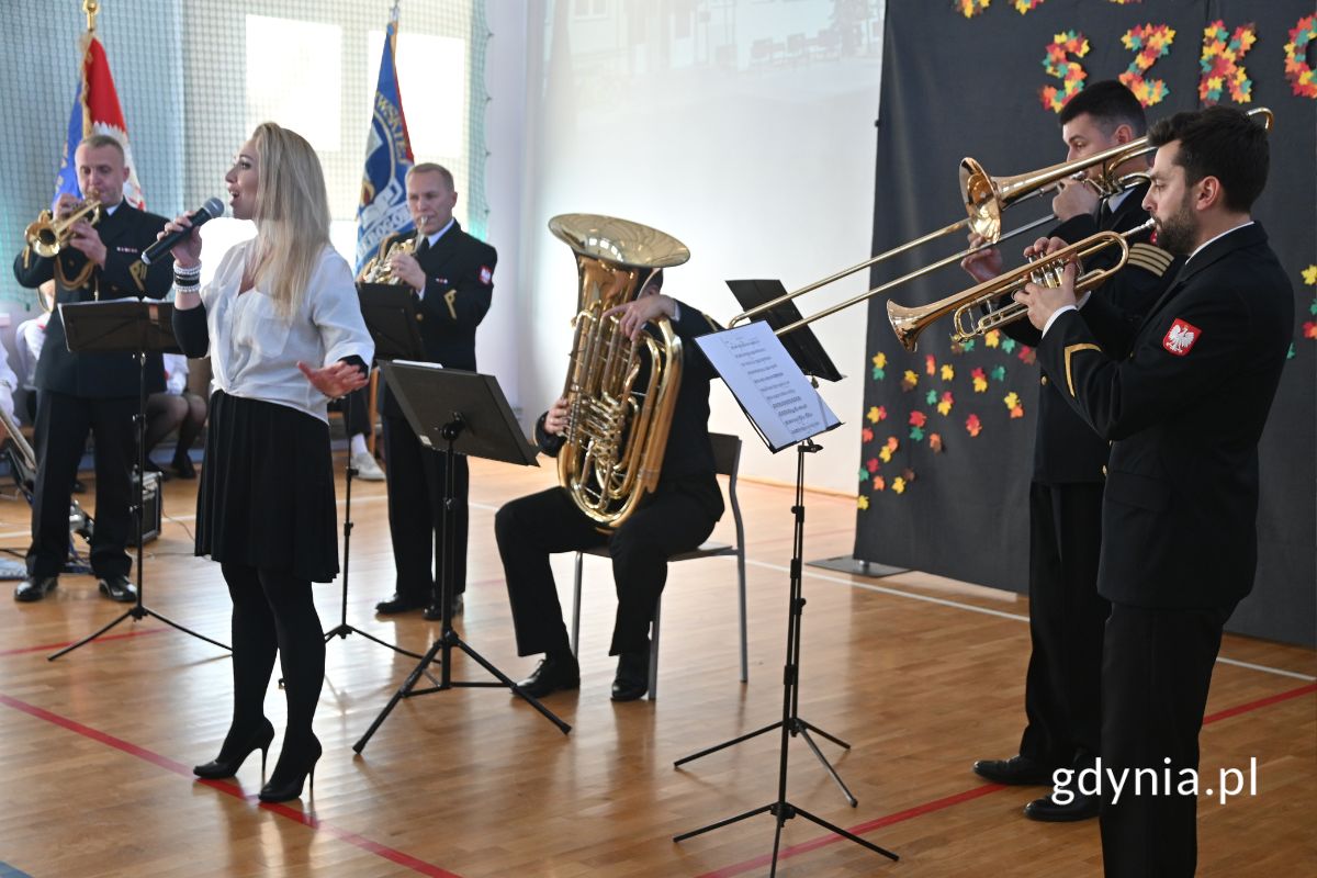 Kobieta śpiewa przy akompaniamencie muzyków z instrumentami dętymi, stojąc w szkolnej hali gimnastycznej