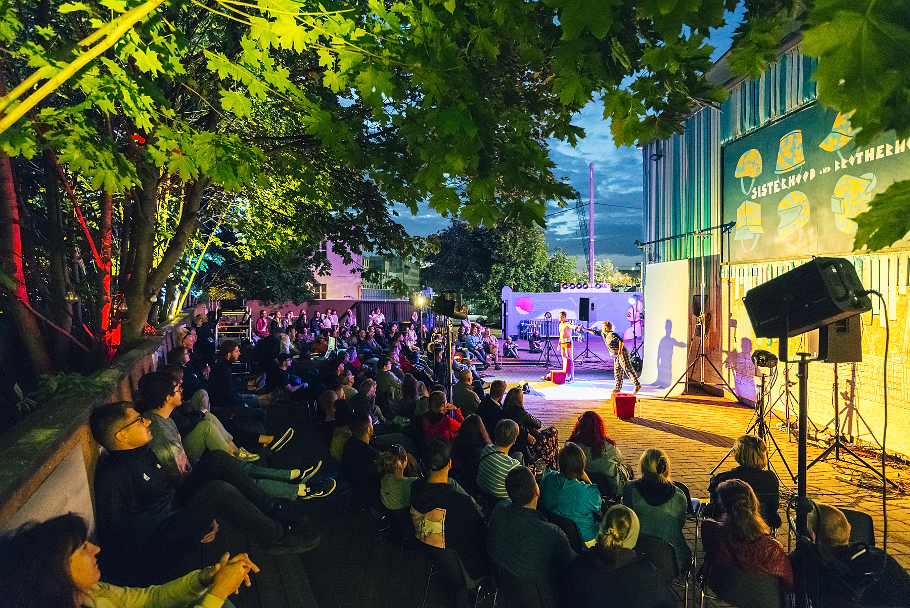 Festiwal &bdquo;Pociąg do Miasta&rdquo; zainaugurował m.in. pokaz spektaklu &bdquo;Mury krzyczą albo uliczna śpiewka&rdquo; Nieformalnego Kolektywu Czy, kt&oacute;ry odbył się na patio Podw&oacute;rka Art przy Klubie Muzycznym "Ucho". Na zdjęciu widać scenę i występujących na niej aktor&oacute;w. Przed sceną i obok niej siedzą widzowie, kt&oacute;ry oglądają przedstawienie // fot. Krzysztof Winciorek