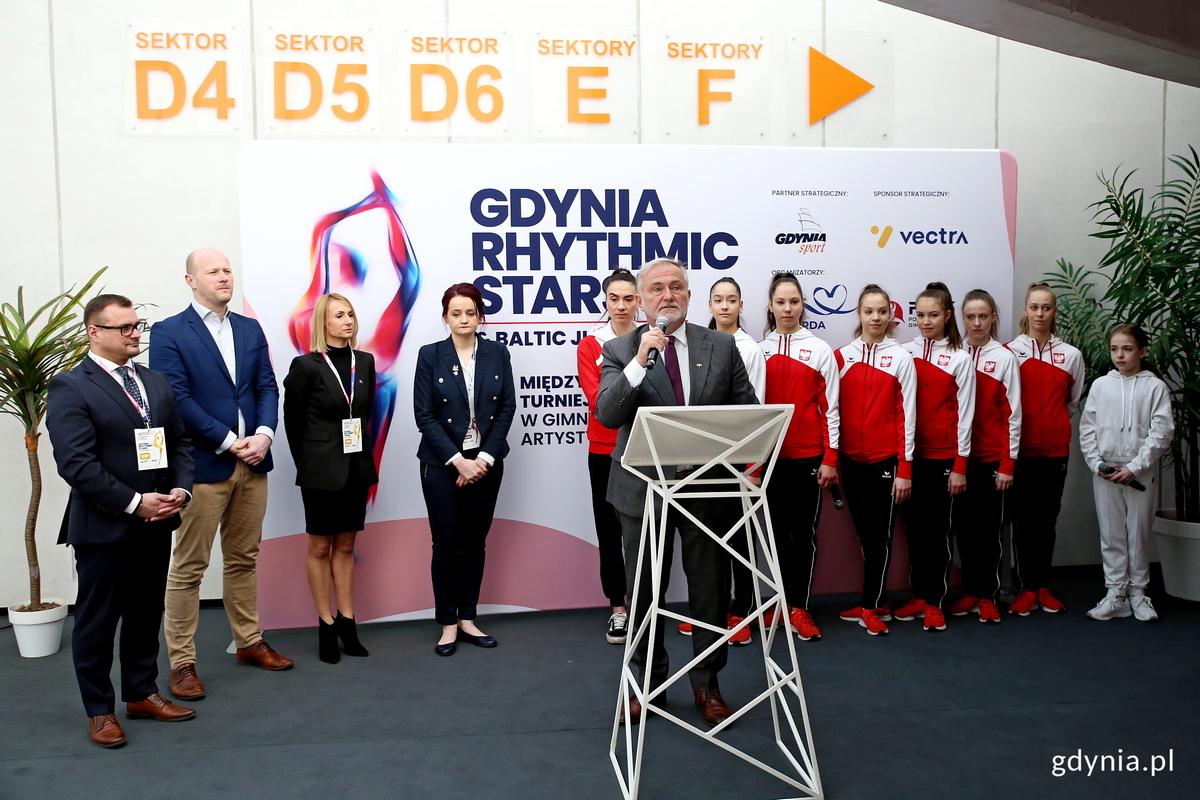 Na zdjęciu widzimy na pierwszym planie prezydenta Gdyni Wojciecha Szczurka. Za jego plecami znajdują się gimnastyczki reprezentacji Polski, wiceprezydent Gdyni Bartosz Bartoszewicz, dyrektorka zawod&oacute;w Anna Mrozińska oraz prezes Polskiego Związku Gimnastycznego Leszek Blanik