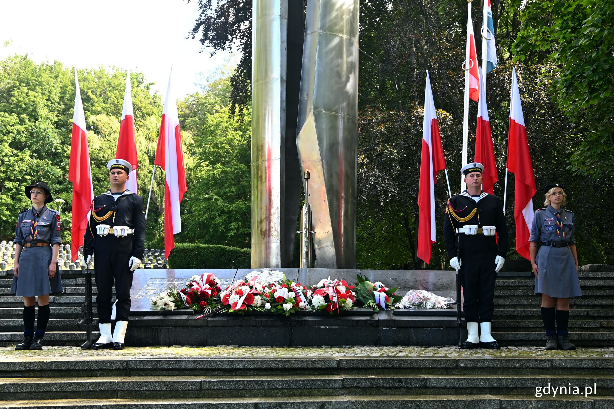 Posterunek honorowy przed pomnikiem Obrońc&oacute;w Wybrzeża na Cmentarzu Wojennym w Redłowie. Po obu stronach pomnika stoją żołnierze i harcerki. W tle biało-czerwonej flagi, zieleń i tablice upamiętniające poległych // fot. Magdalena Czernek