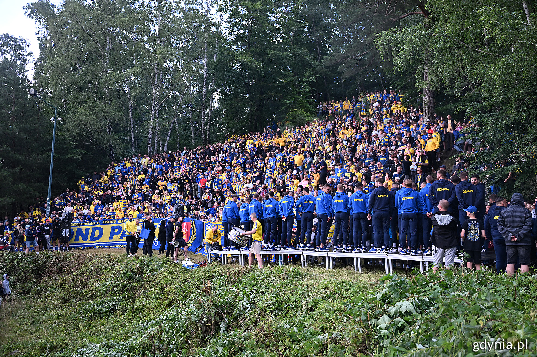 G&oacute;rka przy Ejsmonda, las, skarpa pełna kibic&oacute;w w ż&oacute;łto-niebieskich barwach, na platformie ustawieni piłkarze i sztab szkoleniowy Arki