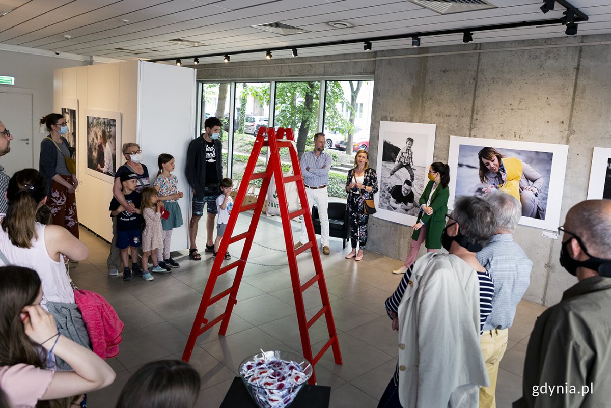 Wernisaż wystawy "Bliskość. Czułość" w Galerii Gdyńskiego Centrum Filmowego. Na środku stoi czerwona drabina. wok&oacute;ł niej stoją uczestnicy wernisażu. Przy zdjęciach na betonowej ścianie: fotograf Piotr Manasterski, Beata Nawrocka - naczelnik Wydziału Polityki Rodzinnej Urzędu Miasta Gdyni i fotograficzka Barbara Matwiejczyk // fot. Przemysław Kozłowski