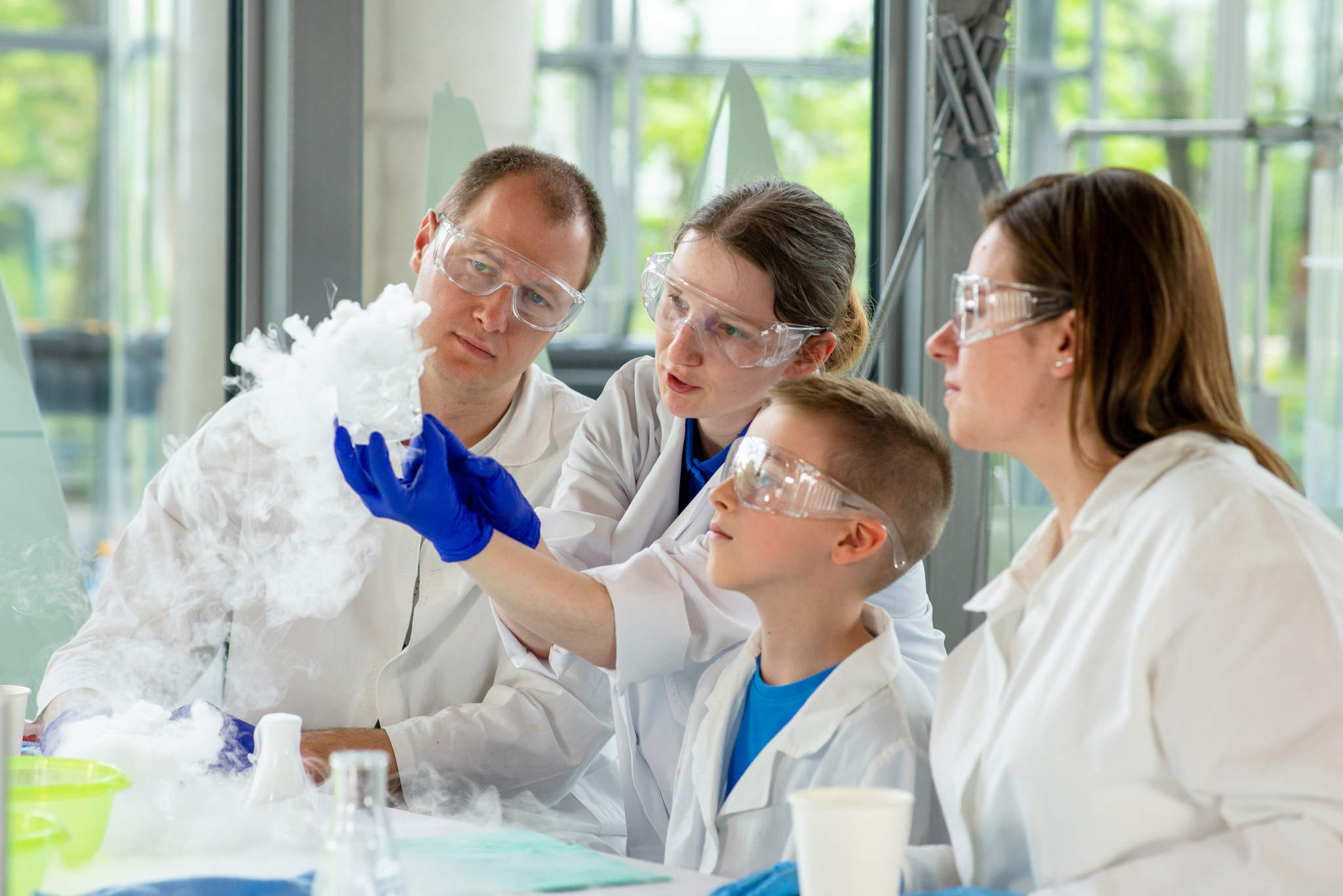 Trzy osoby dorosłe i jedno dziecko ubrane w okulary ochronne robi eksperyment podczas Warsztat&oacute;w Rodzinnych w Centrum Nauki Experyment. Fot. CNE