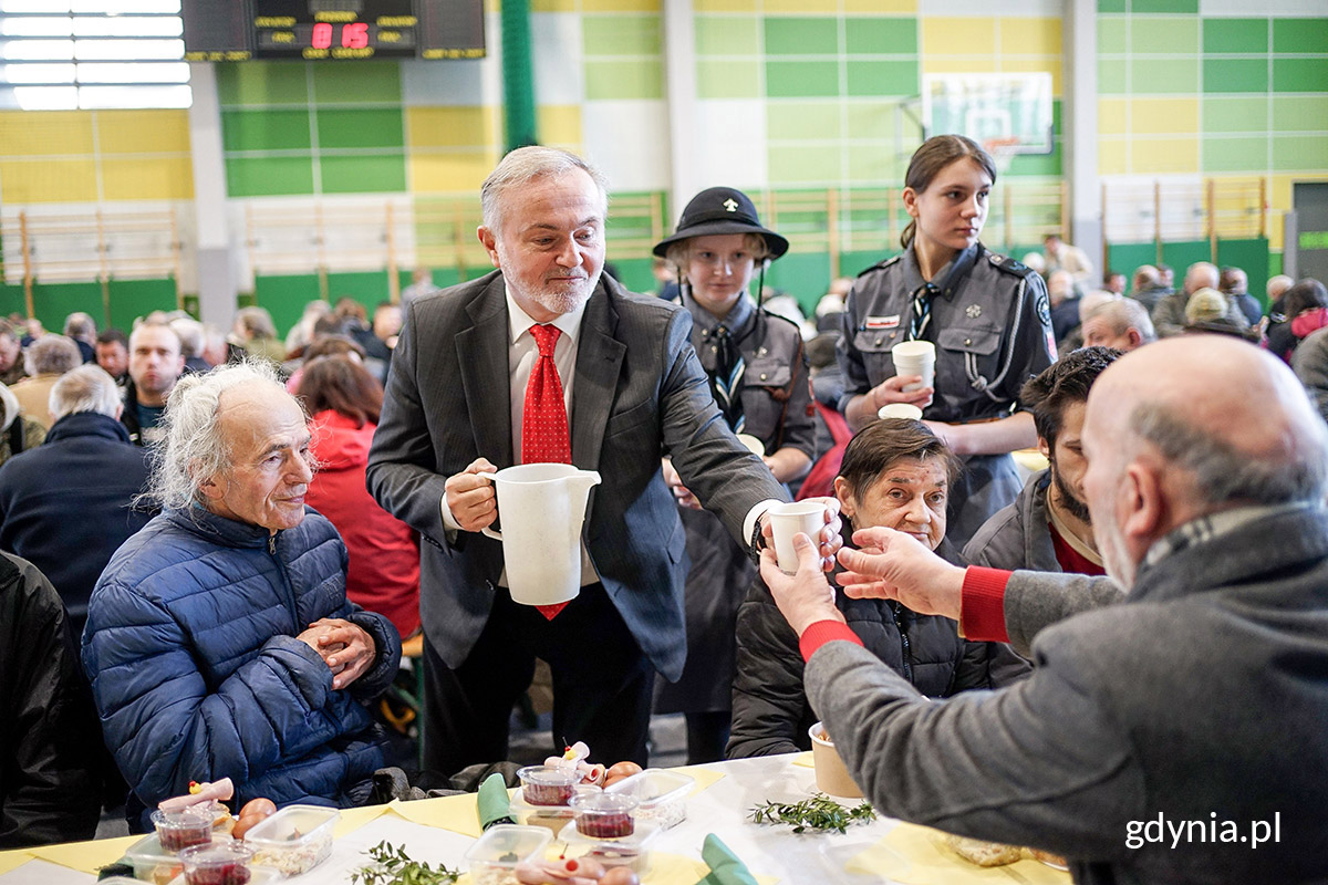 Prezydent Gdyni Wojciech Szczurek nalewa żurek gościom śniadania wielkanocnego dla samotnych i potrzebujących. Obok niego stoją harcerki z kubkami. W tle widać salę gimnastyczną VI LO wypełnioną gośćmi // fot. Mirosław Pieślak