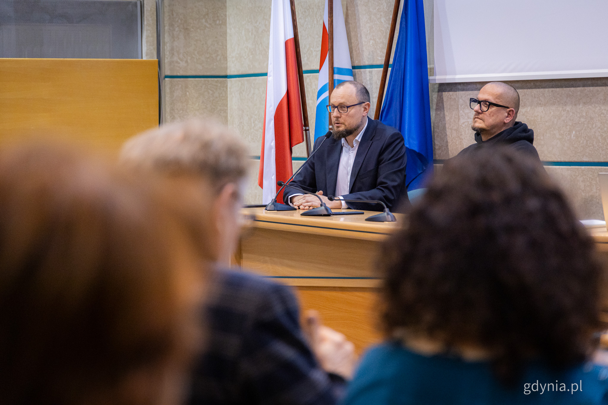 Sala obrad Rady Miasta, flagi miasta, przy stole siedzi Mariusz Bzdęga, widoczni ludzie