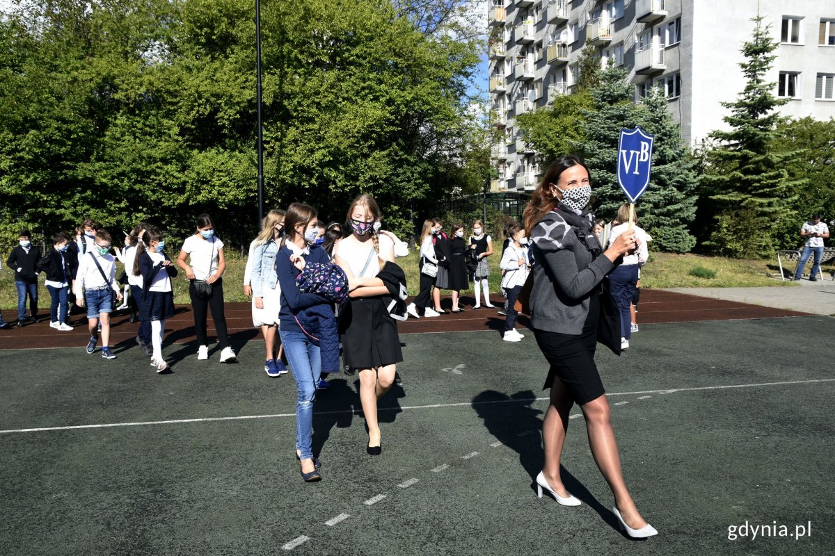 Rozpoczęcie roku w jednej z gdyńskich szk&oacute;ł. Nauczycielka trzyma w ręku niebieską tarczę z nazwą klasy. Za nią w dwuszeregu i odstępach idą uczniowie z maseczkami na twarzach // fot. Magdalena Czernek