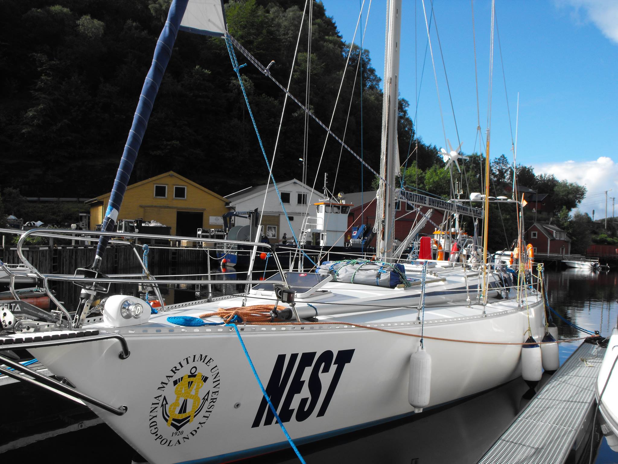 Jacht "Nest" przycumowany do nabrzeża. Na białym kadłubie znajduje się logo Uniwersytetu Morskiego oraz nazwa jednostki - Nest. W tle niskie drewniane budynki i drzewa // fot. archiwum Andrzeja Szklarskiego