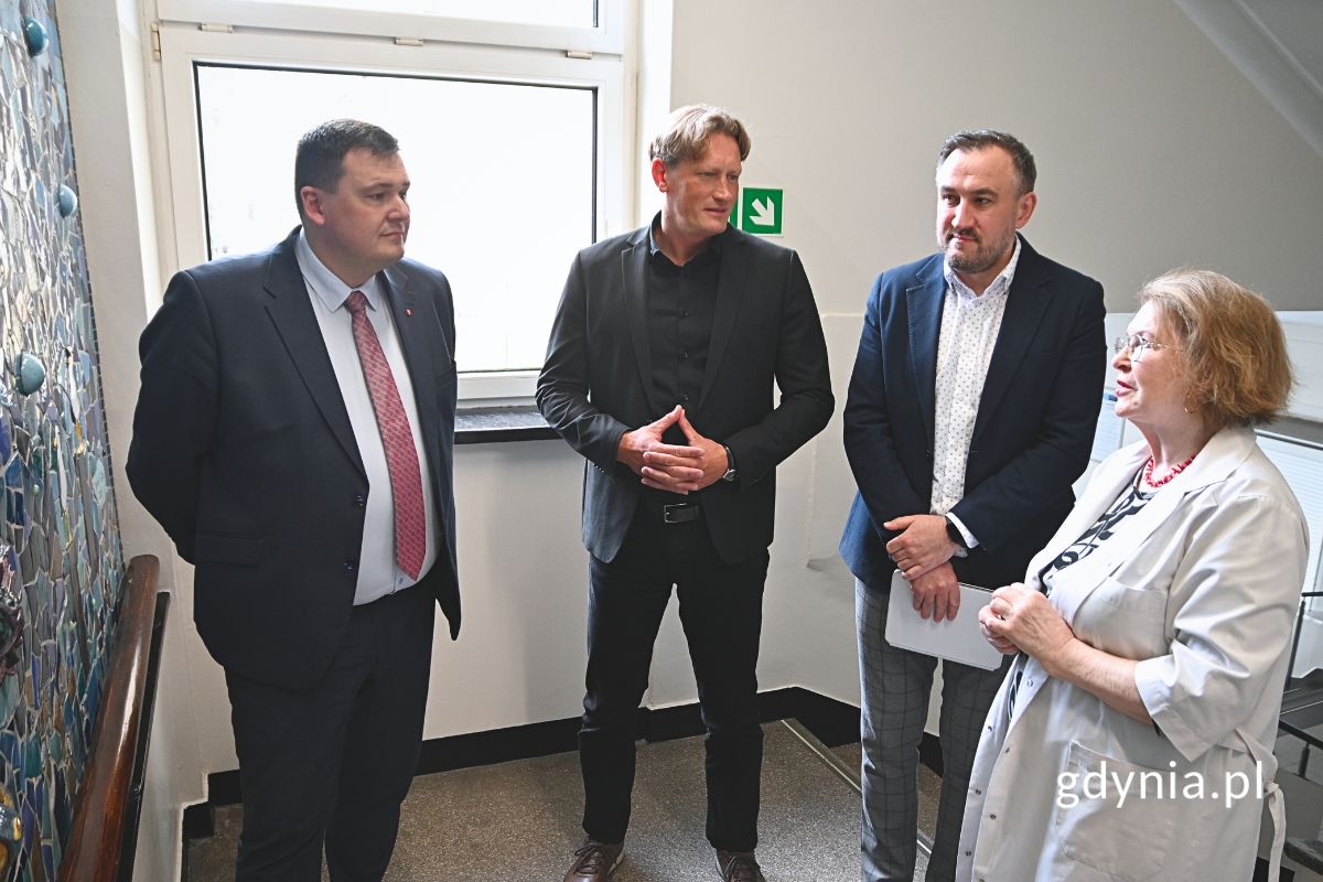 Kobieta w fartuchu lekarskim i trzej elegancko ubrani mężczyźni oglądający kolorową mozaikę na ścianie klatki schodowej