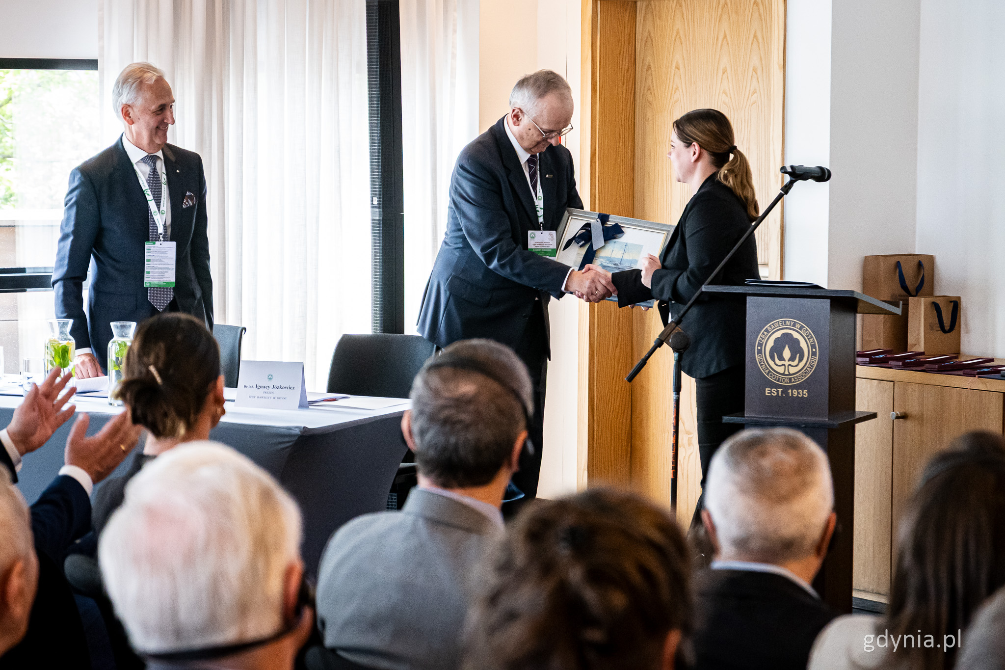 Sala konferencyjna, przy m&oacute;wnicy stoi Aleksandra Kosiorek, przekazuje Ignacemu J&oacute;zkowiczowi prezent w postaci obrazu