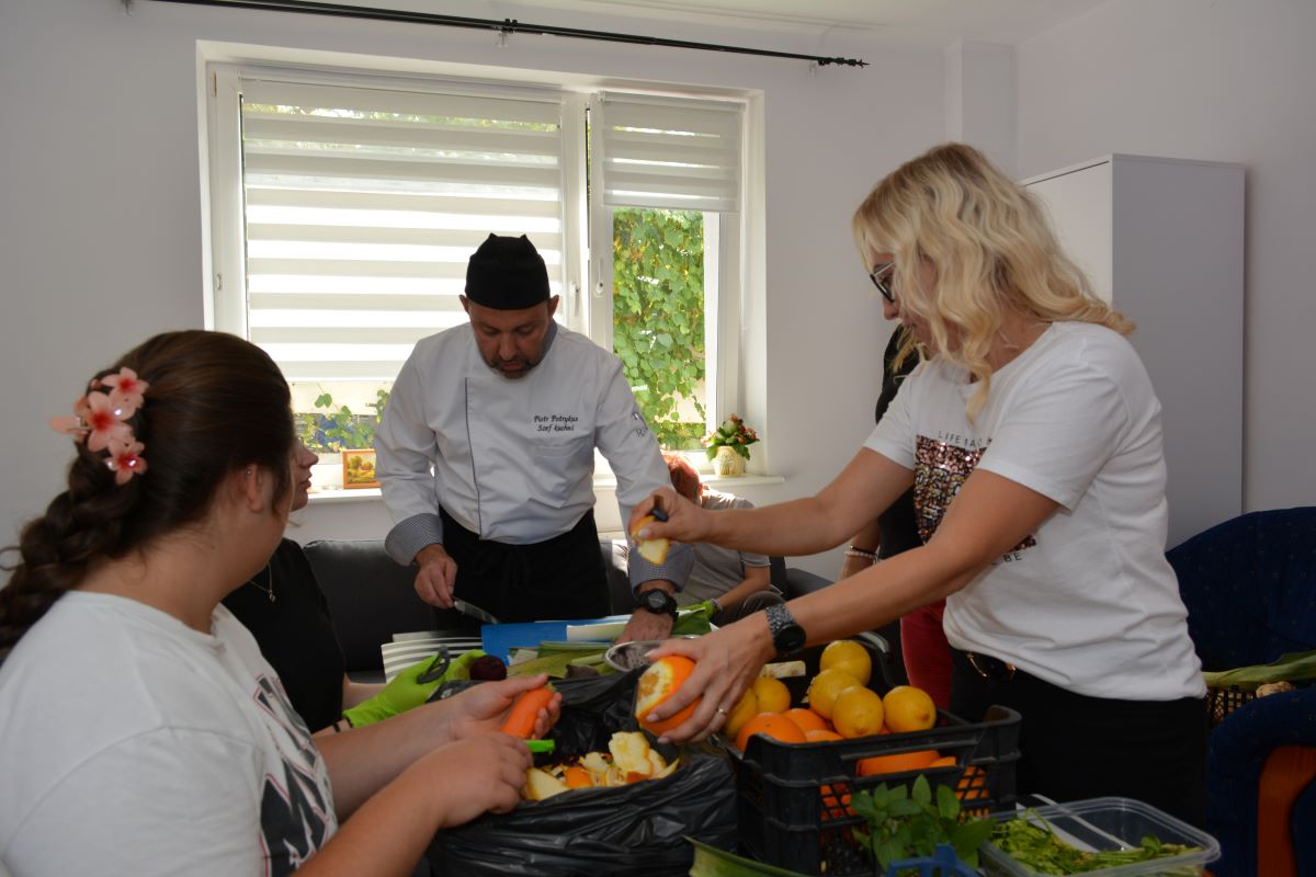 Zdjęcie przedstawia zajęcia poświęcone niemarnowaniu żywności, prowadzone w ramach Gdyńskiego Klubu Mentoringu. Ukazuje grupę młodych ludzi, wraz z profesjonalnym kucharzem po środku. Grupa skupiona jest na dokładnym obieraniu warzyw, z kt&oacute;rych ma powstać p&oacute;źniejszy posiłek. 
