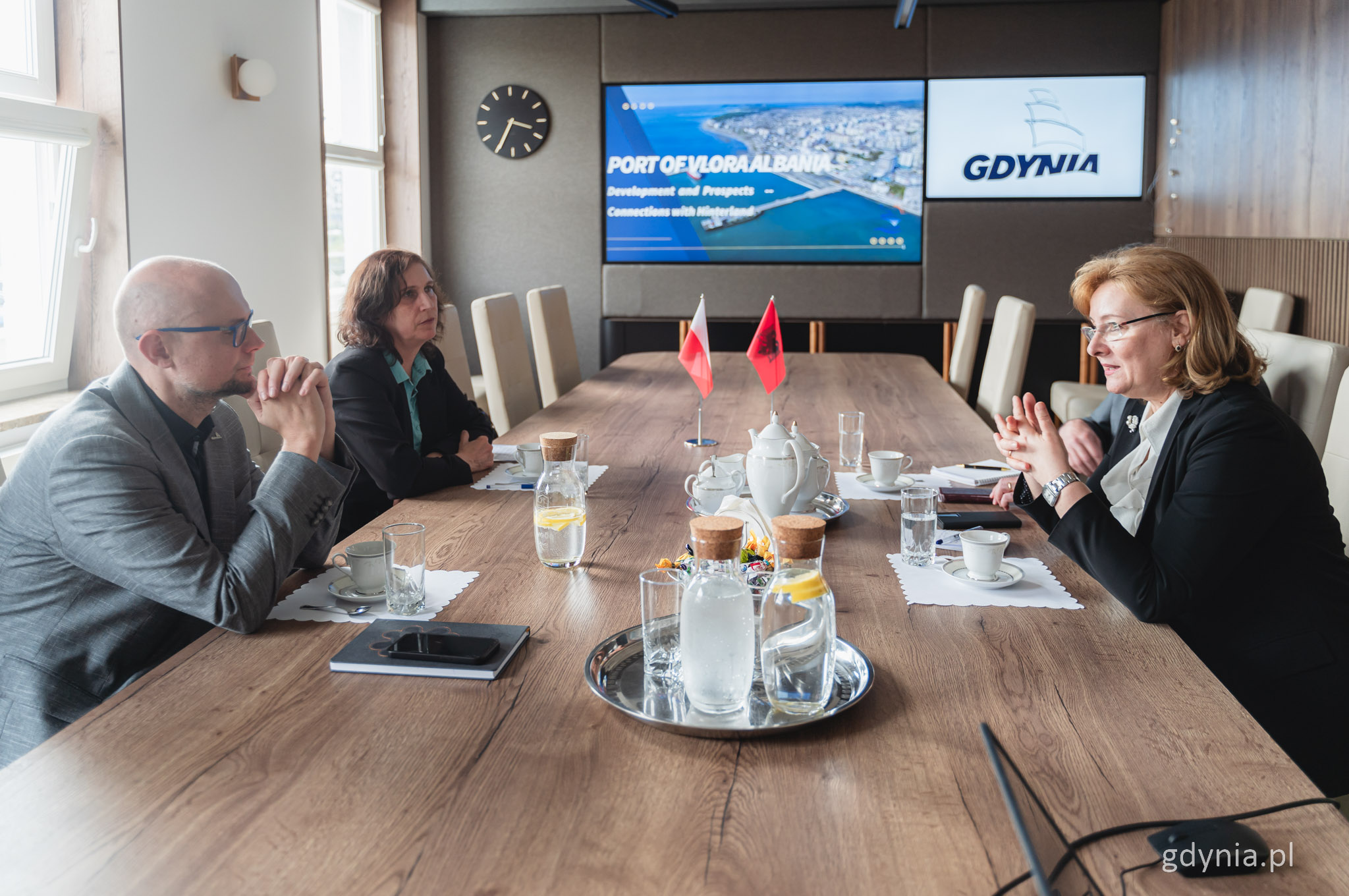 St&oacute;ł w sali konferencyjnej Urzędu Miasta Gdyni, siedzą cztery osoby po obu stronach, na stole napoje i naczynia, w tle wyświetlacze z prezentacją